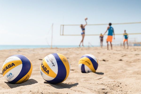 Volleybälle im Sand, dahinter ein Beachvolleyball-Spiel am Meer